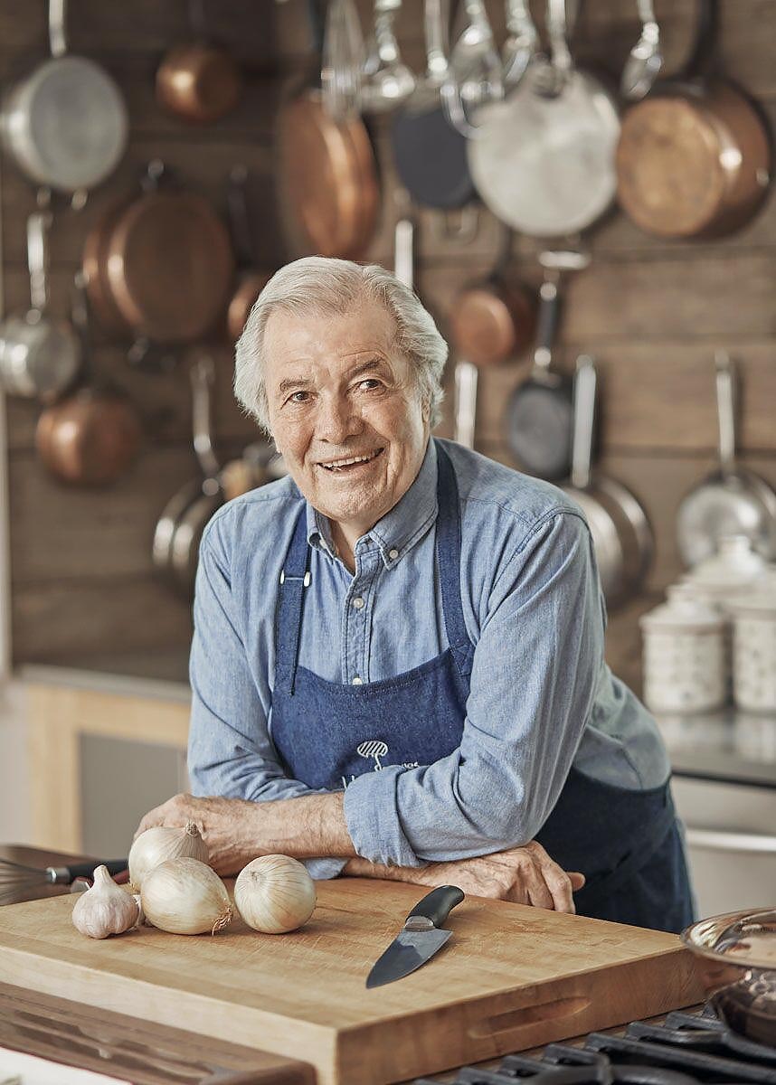 Jacques Pépin in the kitchen with pots hanging behind him